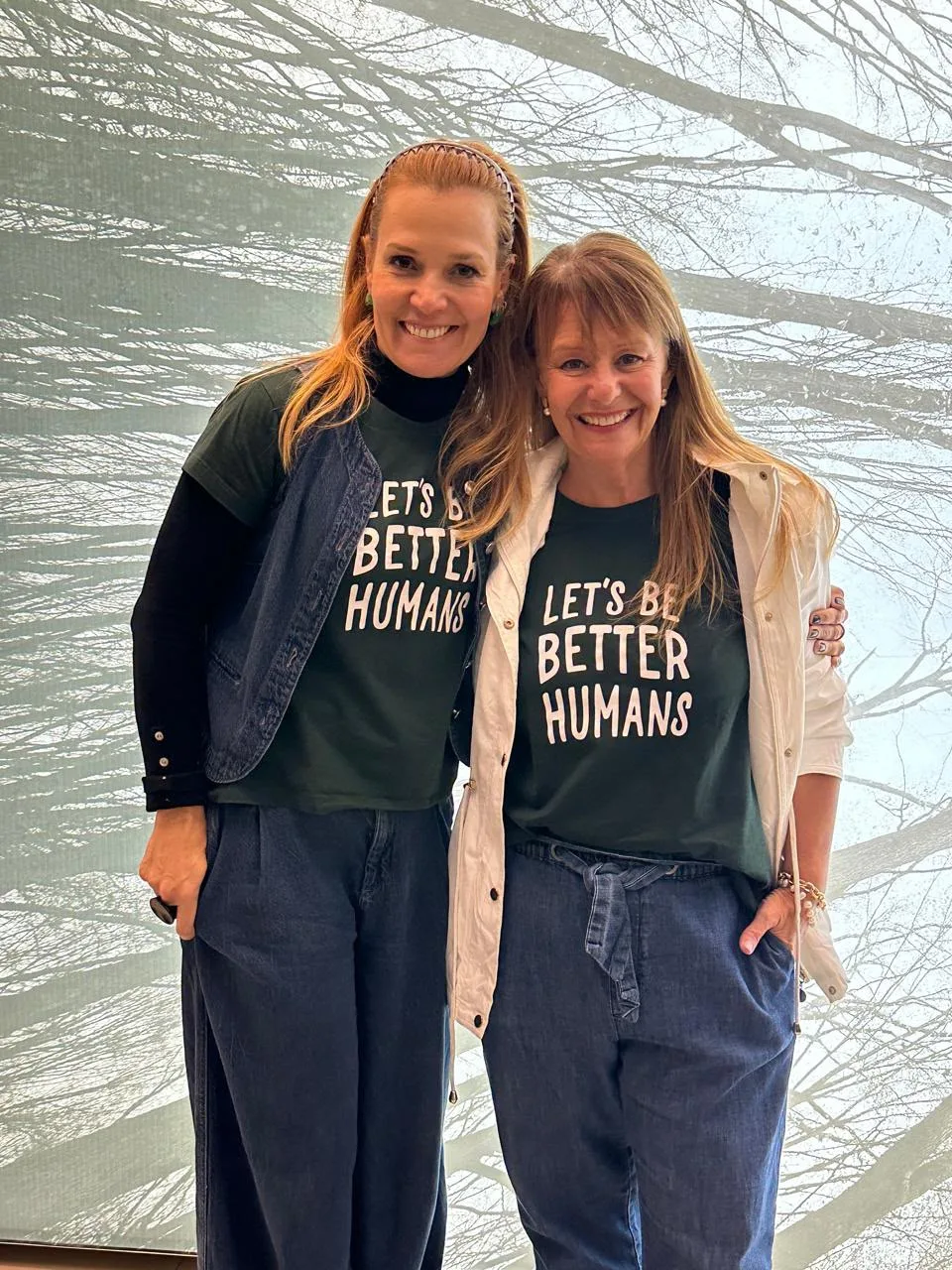 Duas participantes da Confraria Let’s Be posam juntas diante do painel de árvores, de camiseta Let’s Be Better Humans
